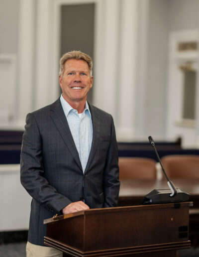 A trustworthy local attorney near me stands behind a wooden podium with a microphone in a courtroom, smiling confidently at the camera. The formal setting features white walls and brown chairs, highlighting his professionalism. -Michael Coulson Law
