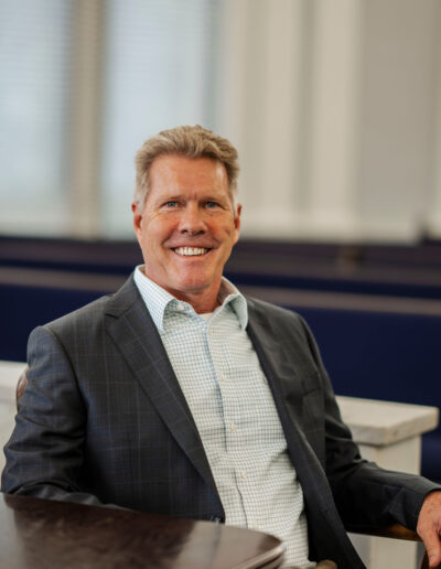 Michael Coulson, wearing a checked shirt and dark suit jacket, is seated indoors, smiling at the camera—embodying the approachable professionalism of a trustworthy local attorney near me. The softly lit background features large windows. -Michael Coulson Law